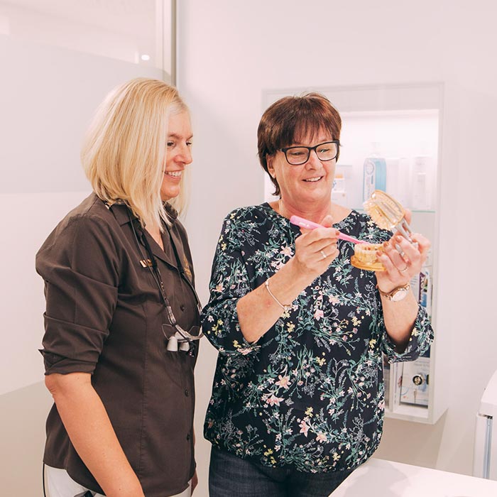 Mitarbeiterin und Patientin reinigen an Gebissmodell Zähne mit Bürste für Prophylaxe beim Zahnarzt in Baden-Baden.