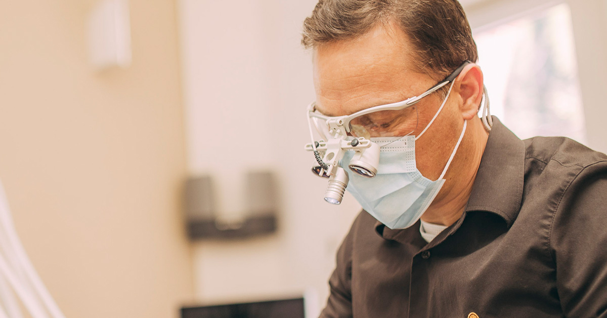 Dr. Thorsten Kamm mit Mundschutz und Lupenbrille: Das Bild zeigt ihn im Halbprofil. Er Blickt konzentriert auf etwas vor sich, dass aber nicht im Bild enthalten ist.