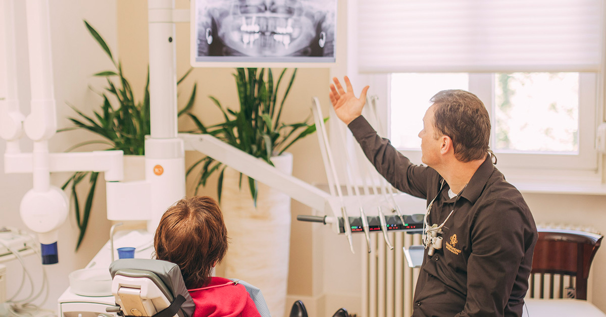 Dr. Torsten Kamm und eine Patientin auf dem Behandlungsstuhl sind von hinten zu sehen. Der Implantologe zeigt der Patienten auf einem Bildschirm das Röntgenbild Ihrer Zähne und bespricht mit ihr das weitere Vorgehen, um die Periimplantitis, eine Entzündung an den Implantaten, in den Griff zu bekommen.